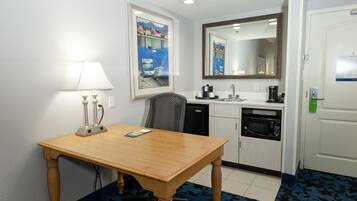 A hotel room with a wooden table, a lamp, a painting, a microwave, and a sink.