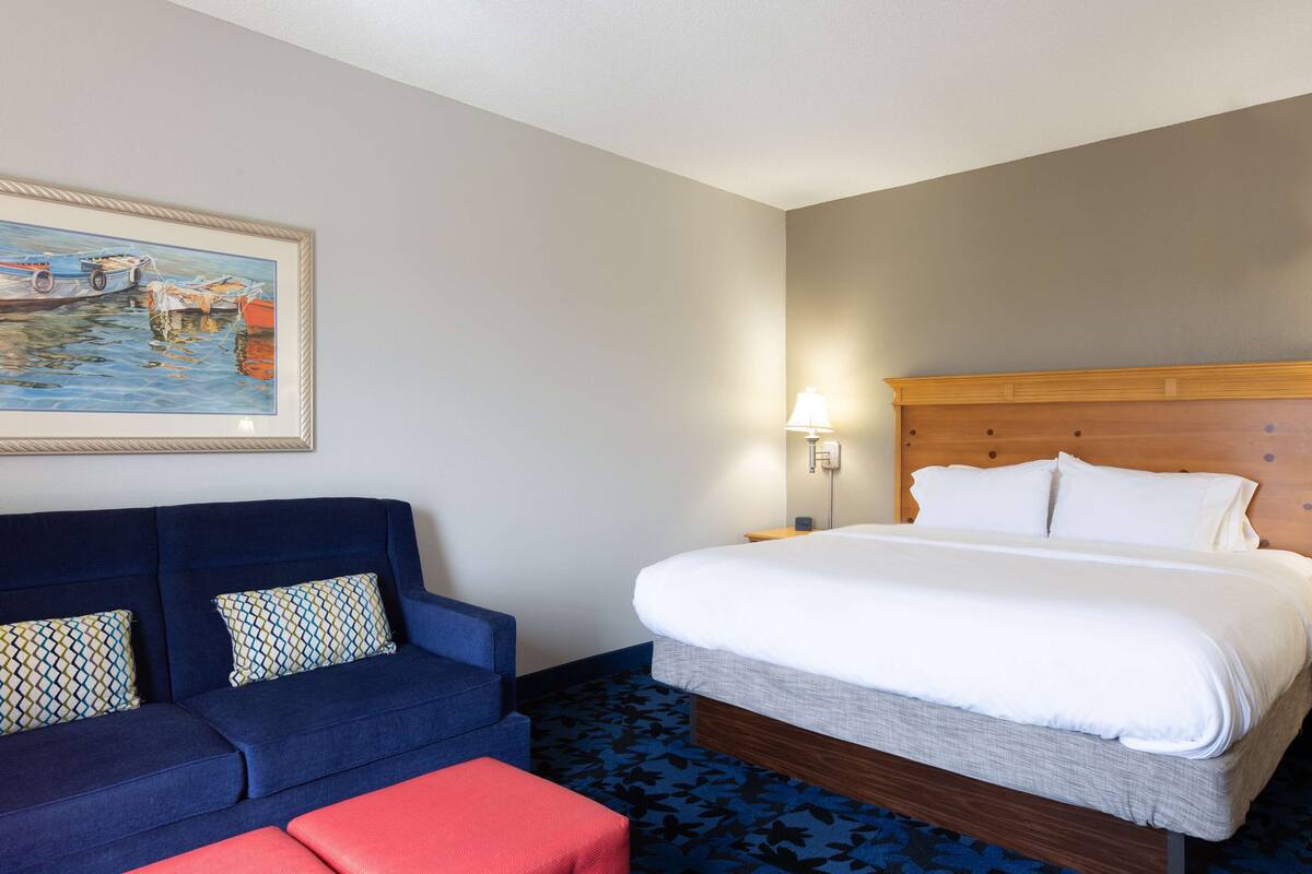 A hotel room with a bed, a blue sofa, a red ottoman, a wall-mounted lamp, and a framed painting of boats.