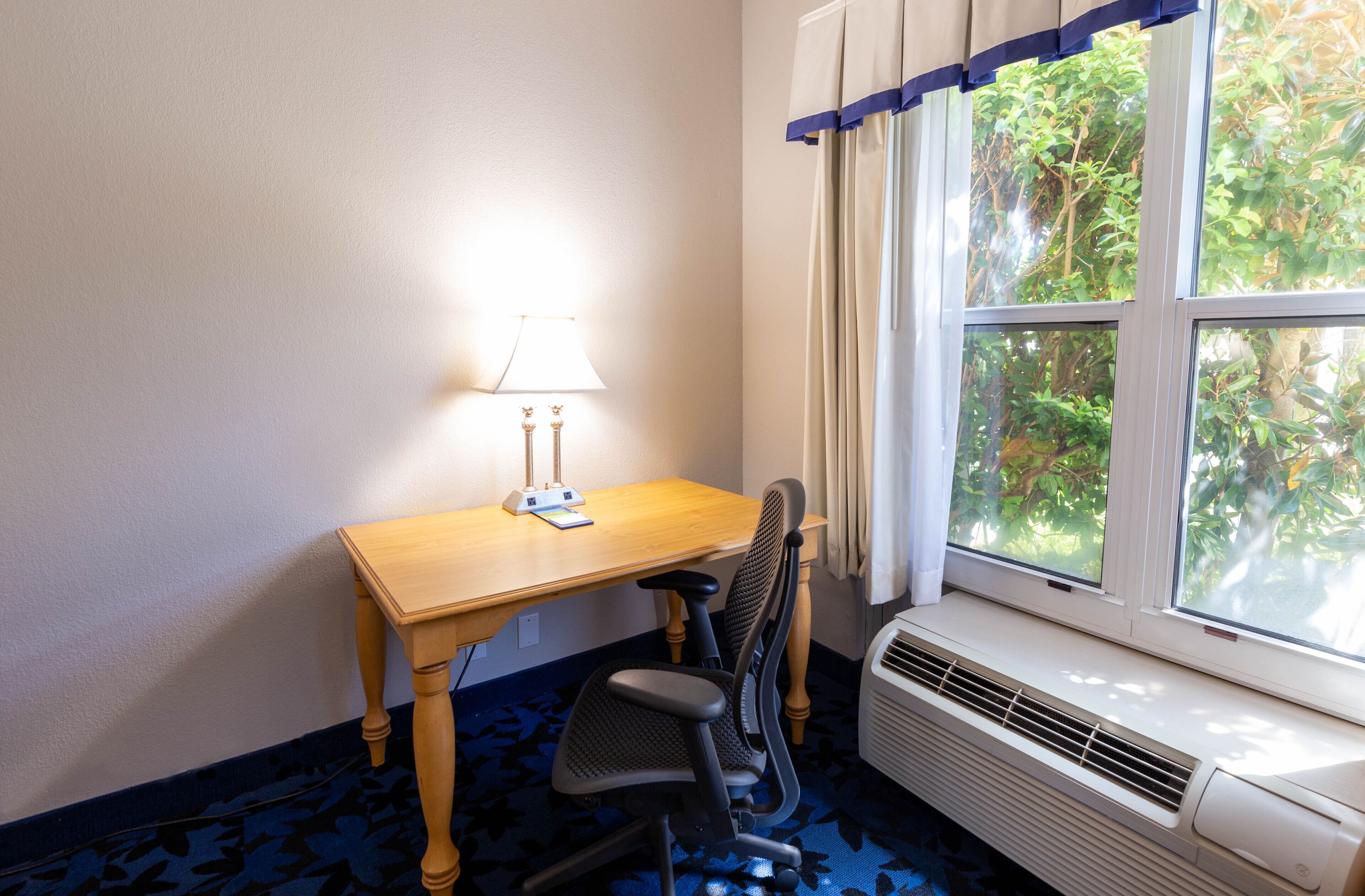 Una habitación con escritorio de madera, una silla, una lámpara y una ventana con cortinas.