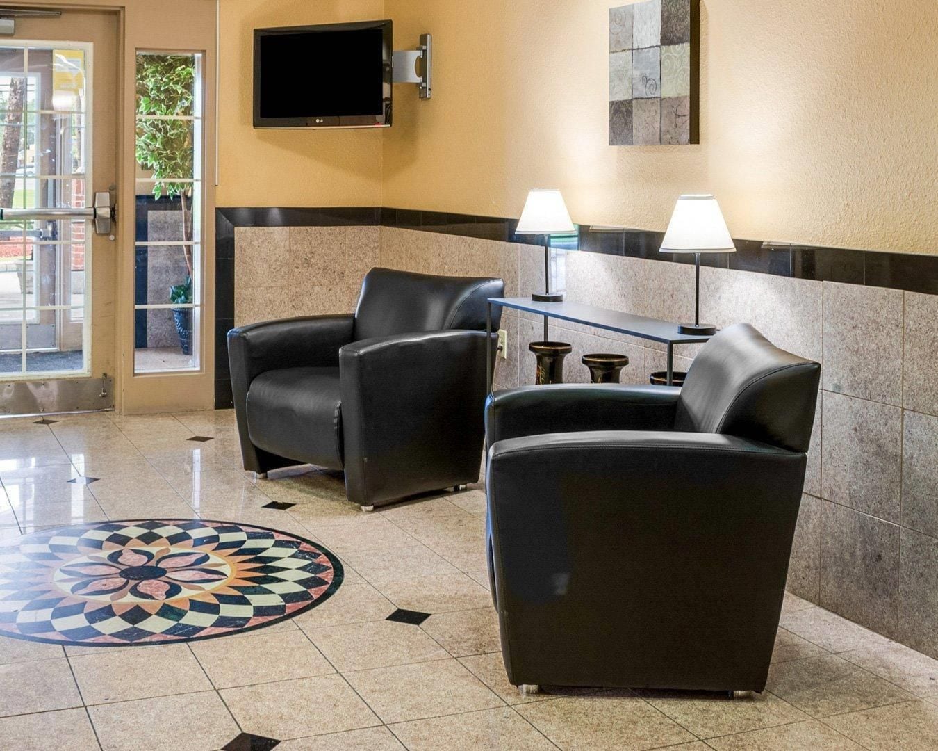 A lobby with two black leather armchairs, a small table with two lamps, a flat-screen TV mounted on the wall, and a patterned floor design.