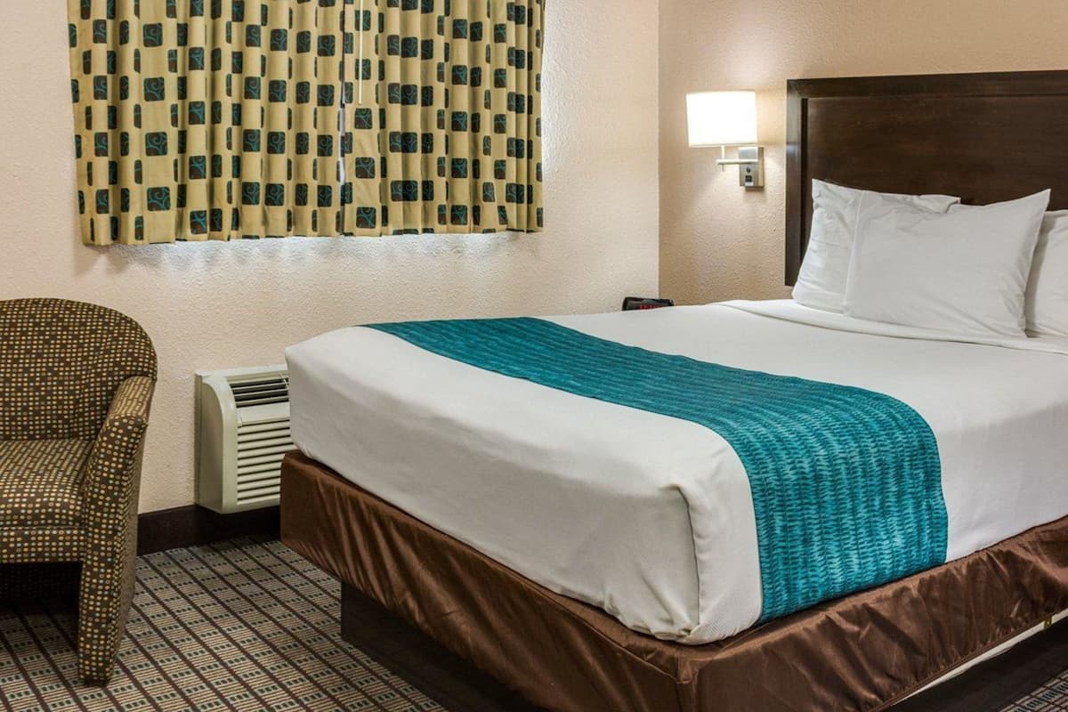 A hotel room with a bed, a chair, a wall-mounted lamp, and a window with patterned curtains.