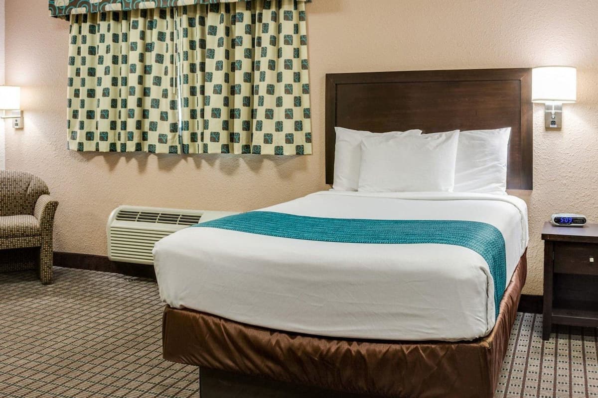 A hotel room with a large bed, a patterned curtain, a chair, a bedside table with a lamp, and a wall clock.