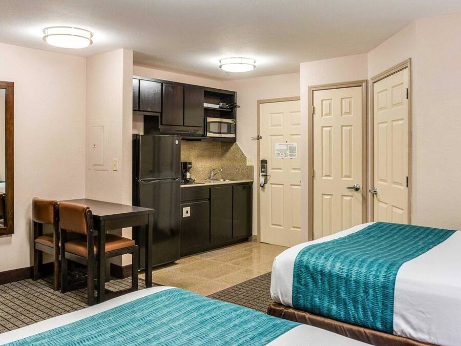 A hotel room with two beds, a kitchenette, a dining table with a chair, and two doors.