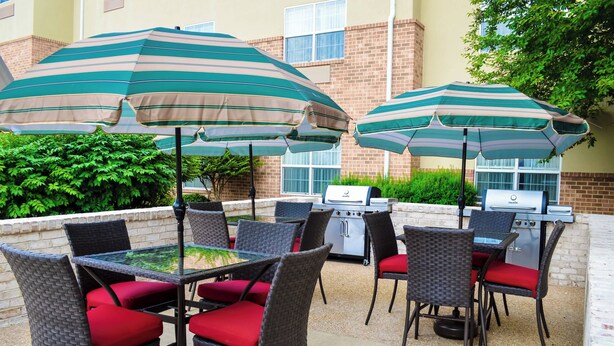 Une terrasse extérieure avec des fauteuils en osier et des coussins rouges, deux parasols à rayures et un barbecue.