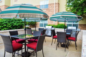 Une terrasse extérieure avec des fauteuils en osier et des coussins rouges, deux parasols à rayures et un barbecue.
