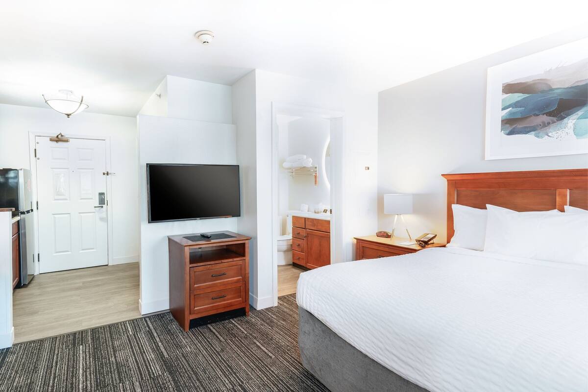 Een hotelkamer met een groot bed, een flatscreen televisie, houten meubels, een badkamer met douche en een schilderij aan de muur.