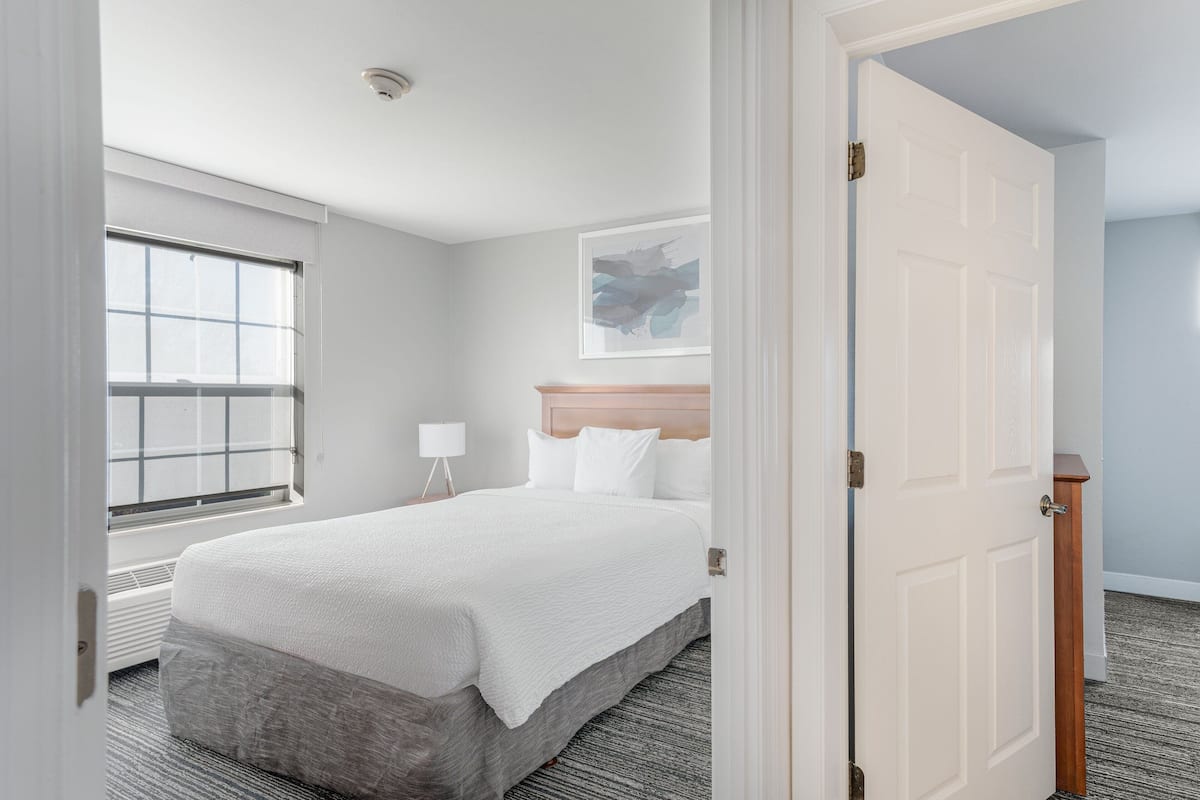 Een hotelkamer met een groot bed, een raam, een bedlamp en een lijst met kunstwerk aan de muur.