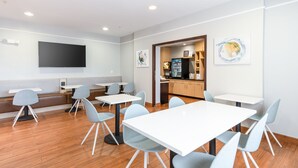 Un espace repas moderne avec des tables blanches et des chaises grises, un téléviseur à écran plat fixé au mur et une cuisine visible à travers une porte ouverte.