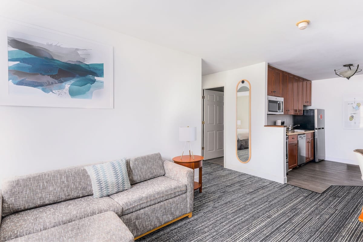 Een moderne woonkamer met een grijze bank, een rond bijzettafeltje en een groot abstract schilderij. Achtergrond toont een keuken met houten kasten en moderne apparatuur.