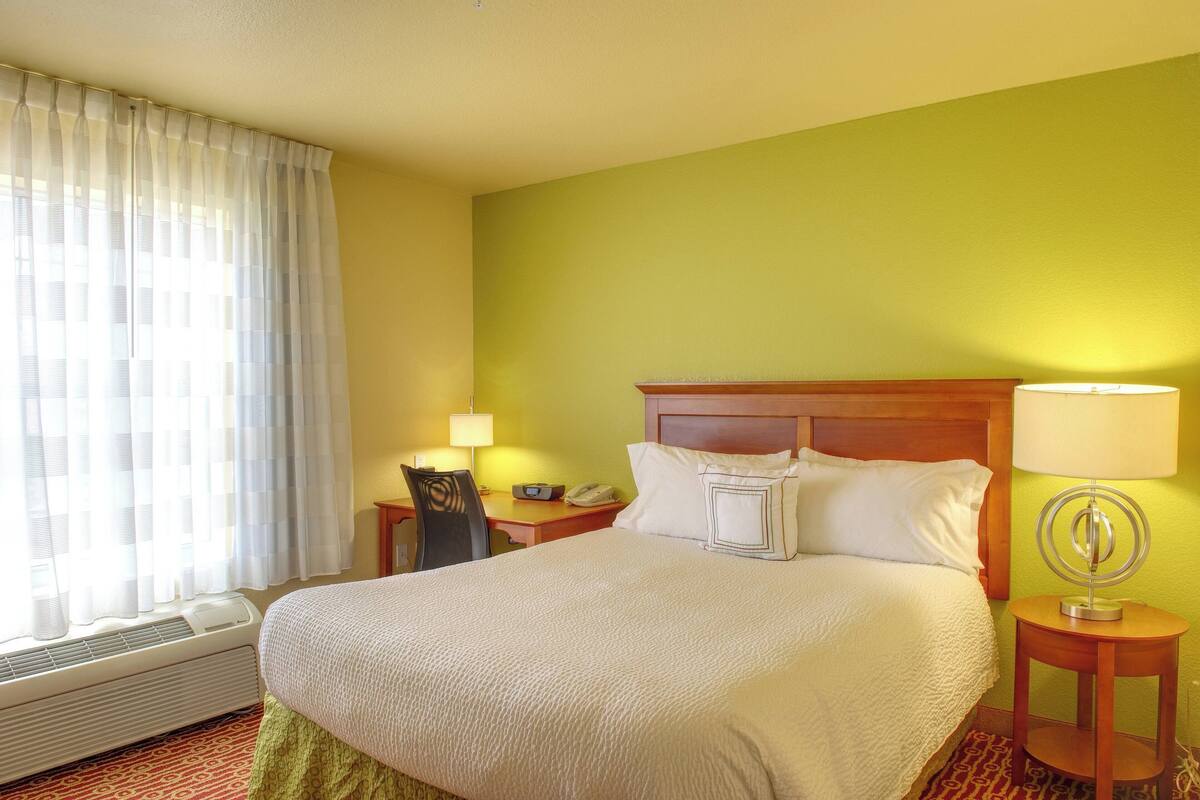 Een hotelkamer met een bed, een bureau, een stoel, een lamp en een raam met gordijnen.