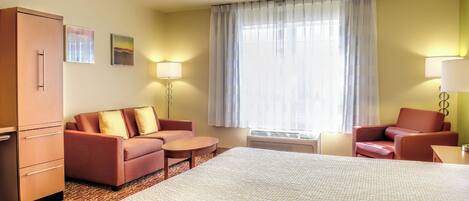 A hotel room with a large bed, two sofas, a small table, a desk, a wardrobe, and a large window with curtains.
