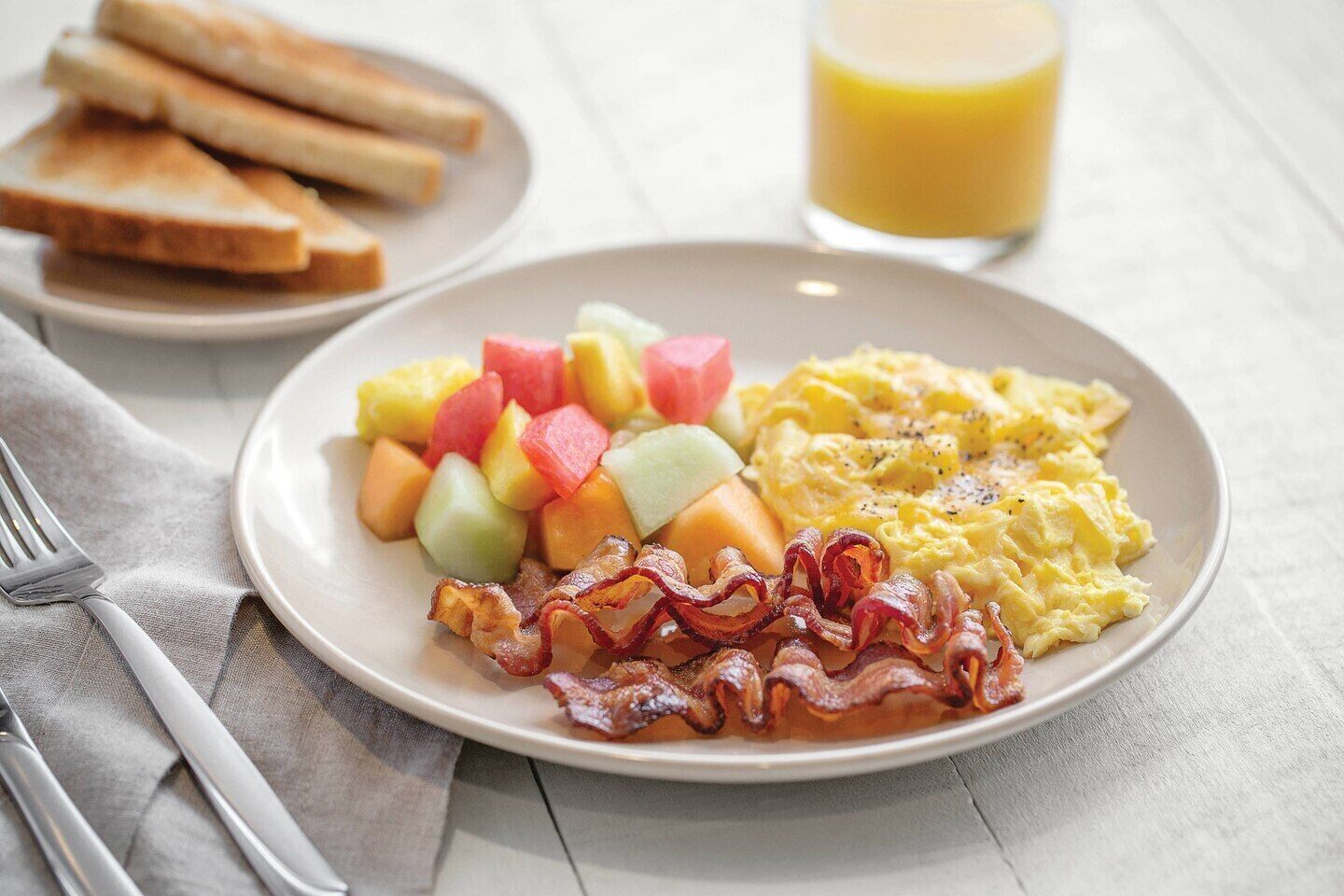 A breakfast plate with scrambled eggs, bacon, and diced fruit, accompanied by toast and a glass of orange juice.