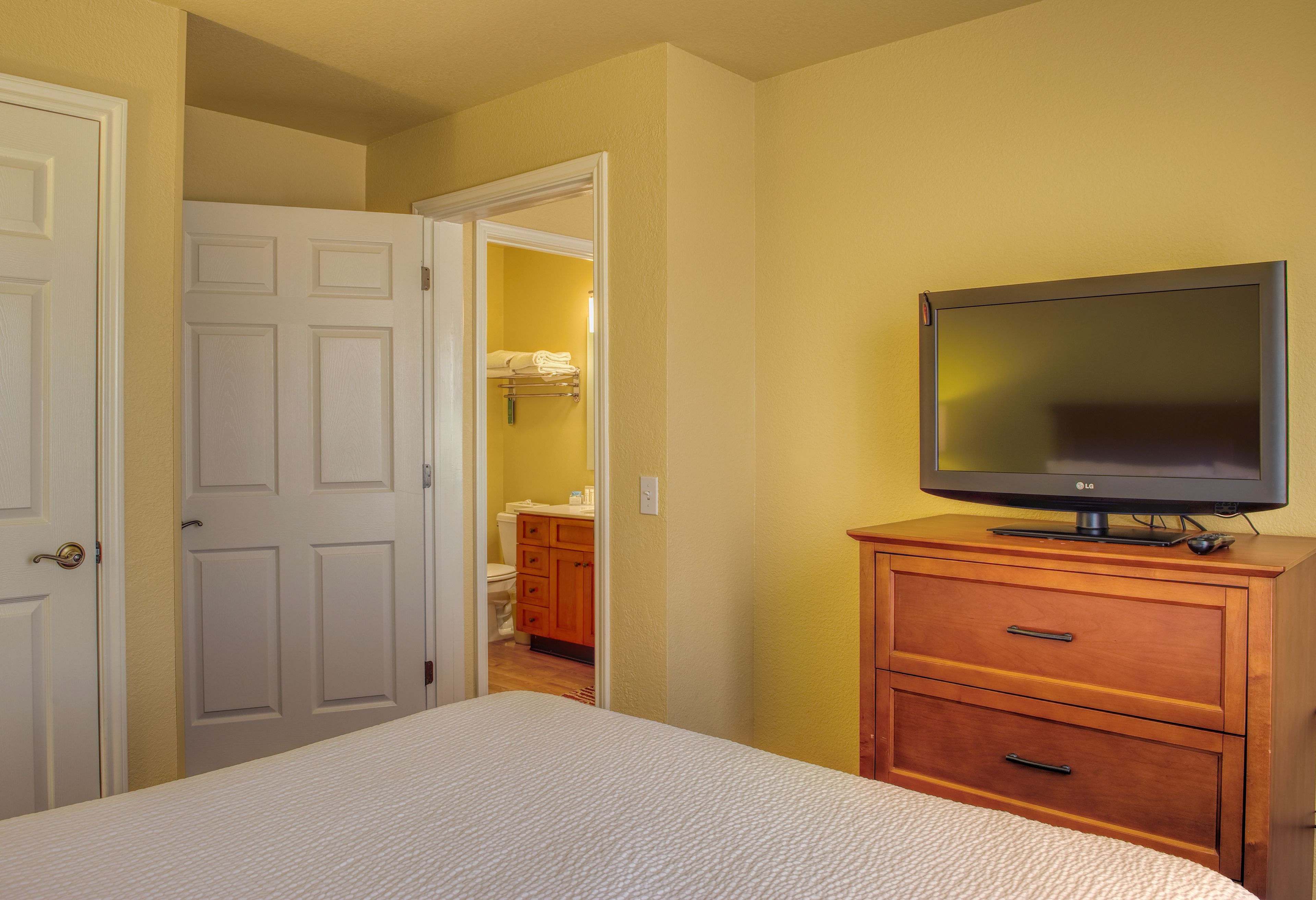 A hotel room with a bed, a wooden dresser, a television, and a bathroom visible through an open door.