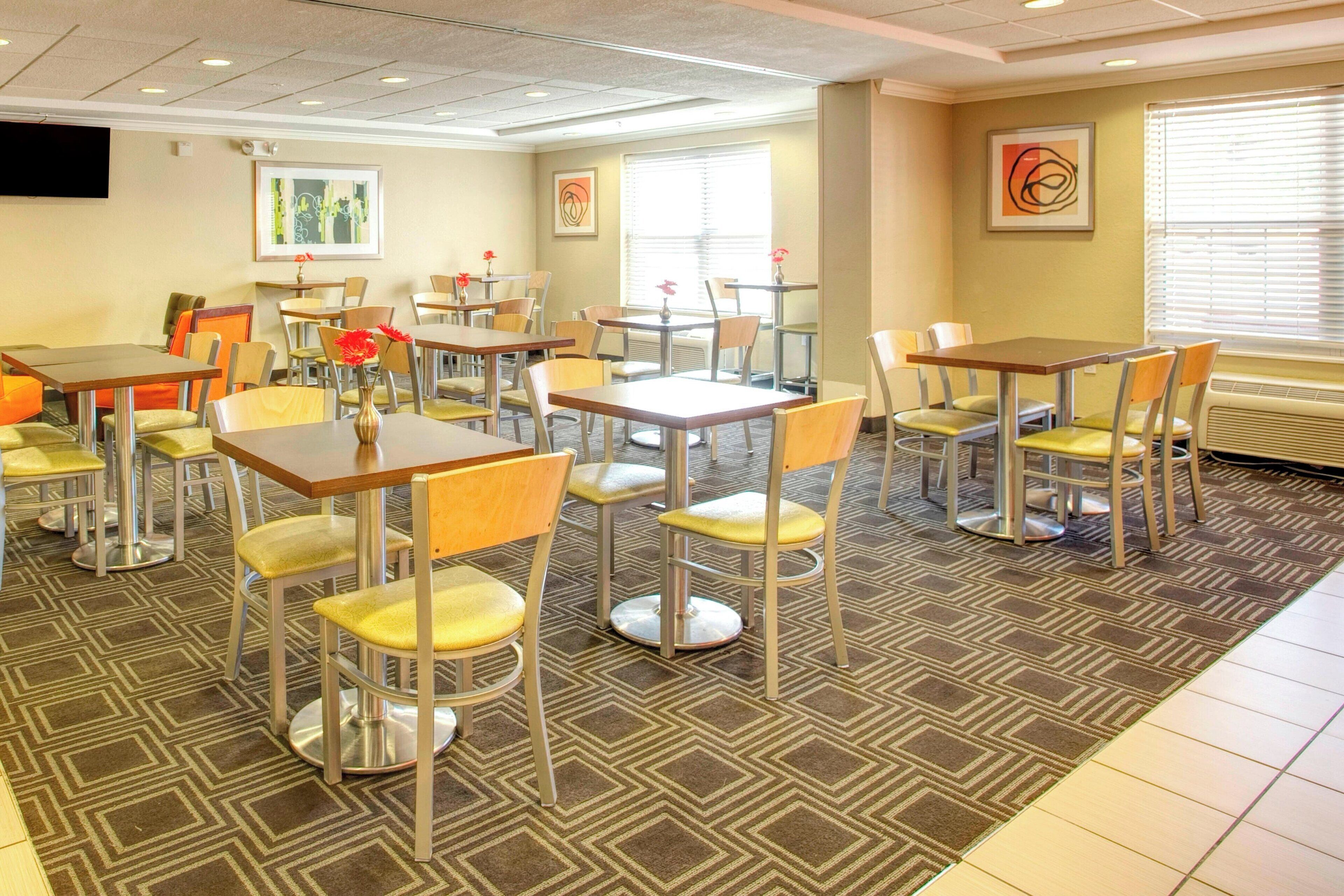 A hotel dining area with multiple tables and chairs, a flat-screen TV mounted on the wall, and artwork on the walls.