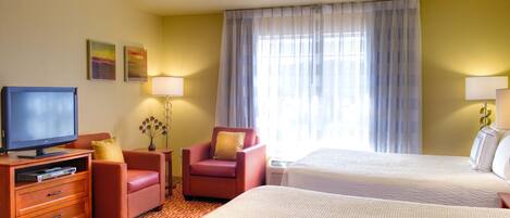 A hotel room with a TV, two red armchairs, a desk, a lamp, and two beds.