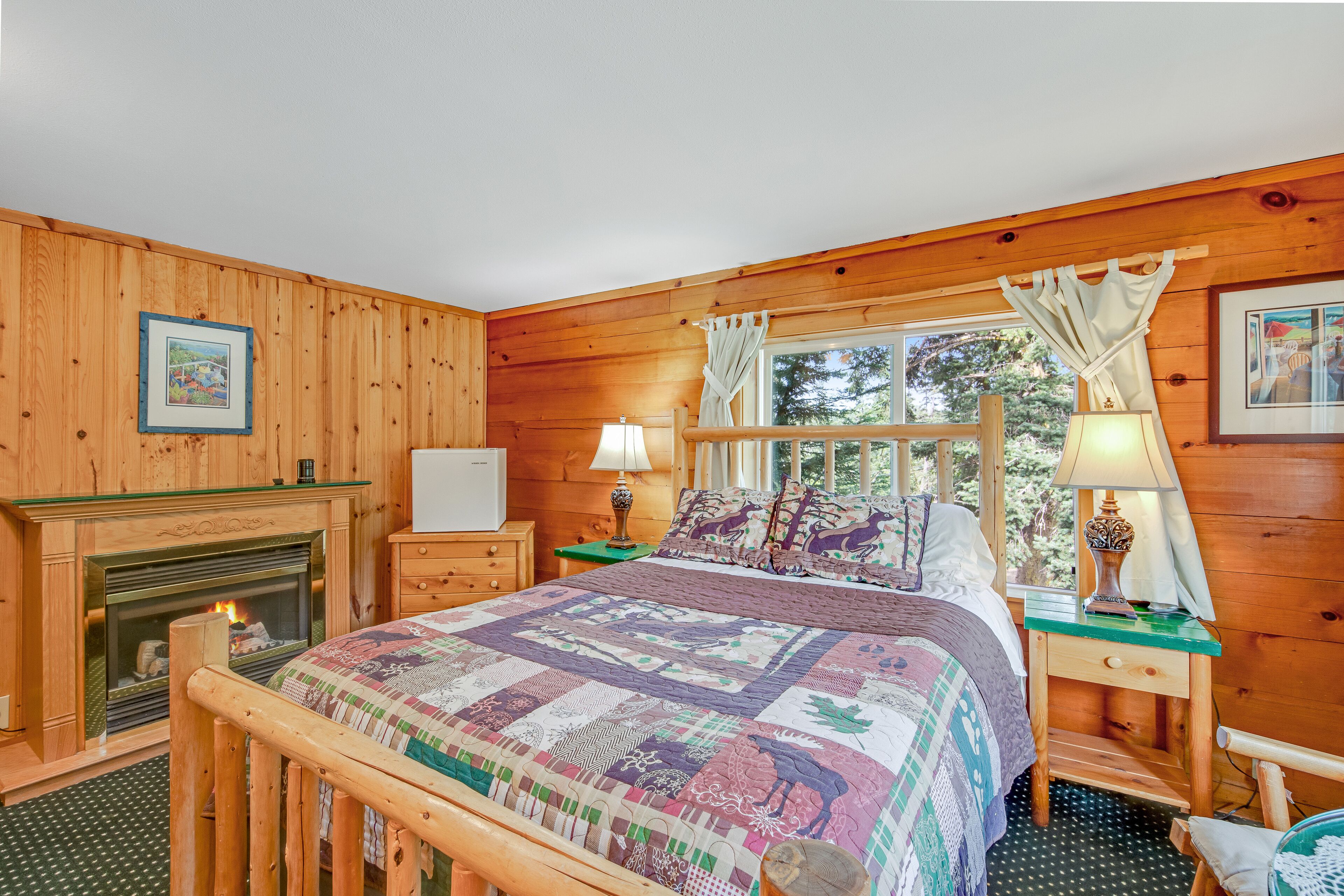 A cozy wooden cabin bedroom with a large bed, a fireplace, and a window with curtains.