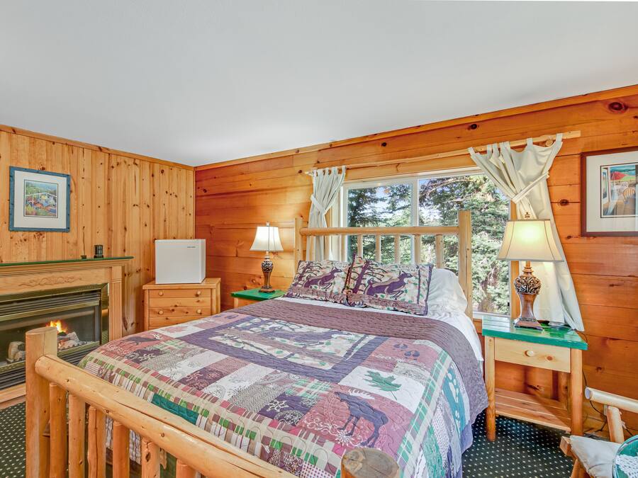 A cozy wooden cabin bedroom with a large bed, a fireplace, and a window with curtains.