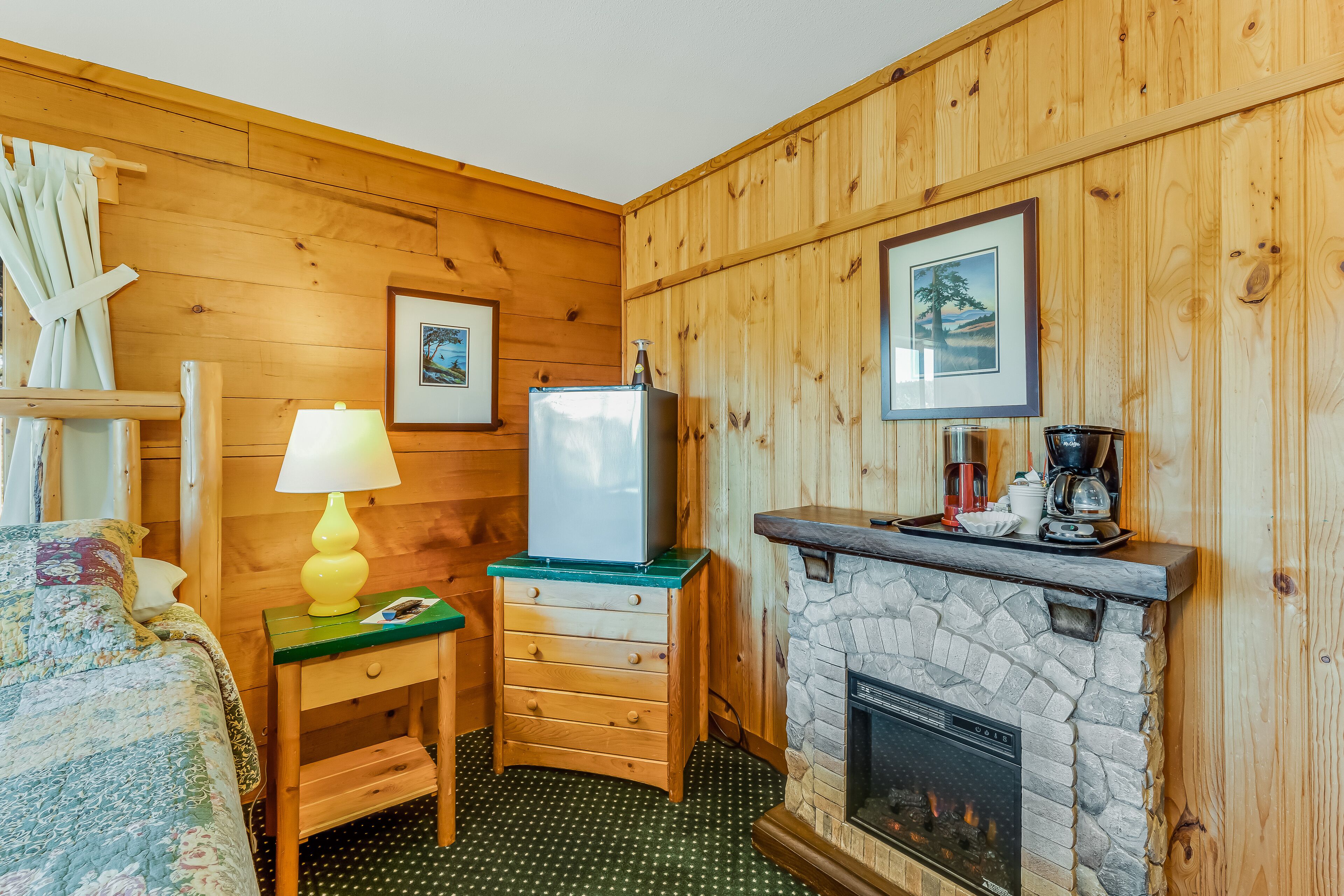 Une pièce confortable avec un lit, une commode, une lampe, un réfrigérateur, une cheminée et des tableaux encadrés accrochés au mur.