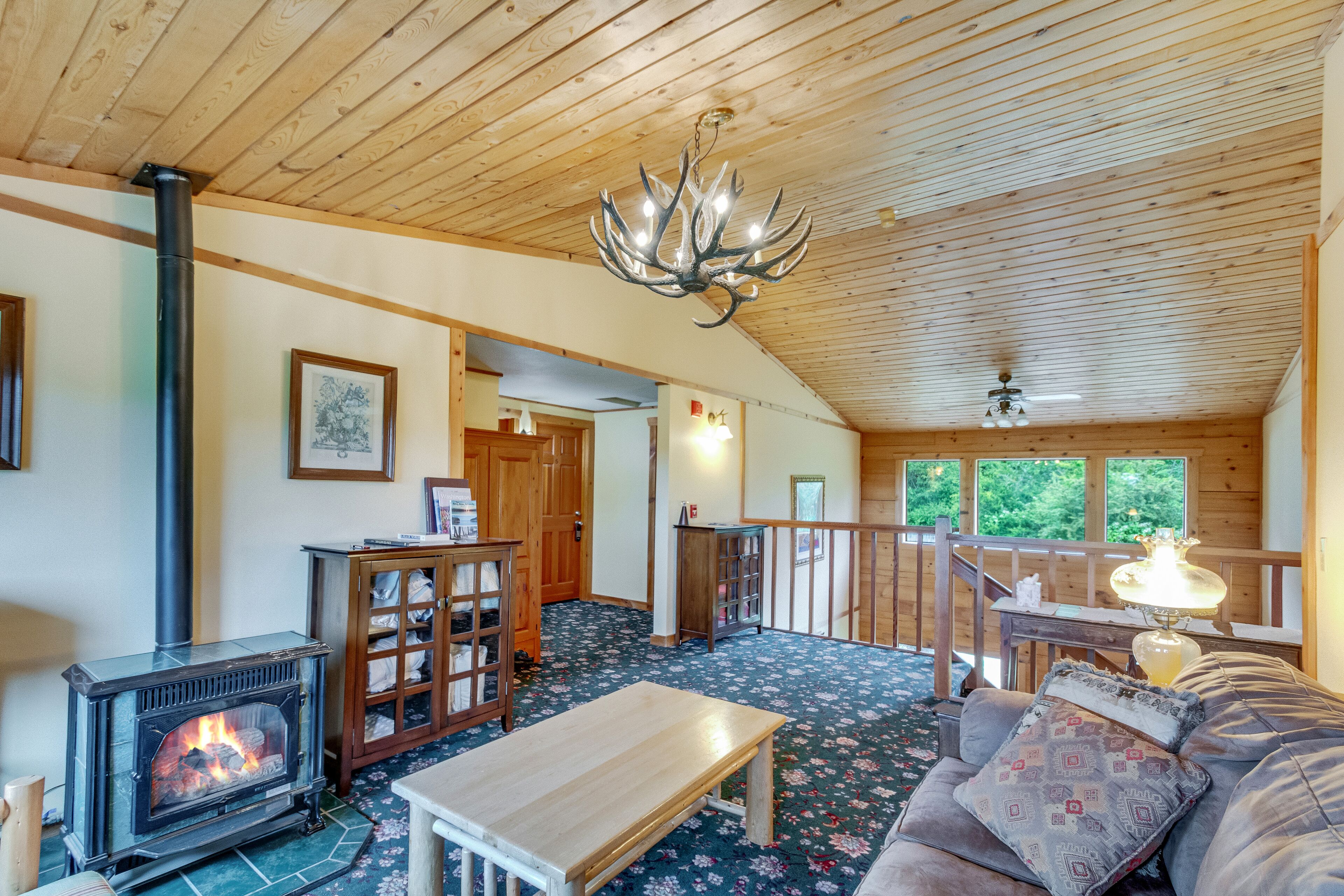 A cozy cabin interior with a wood-burning stove, a cushioned sofa, a wooden table, and a chandelier.