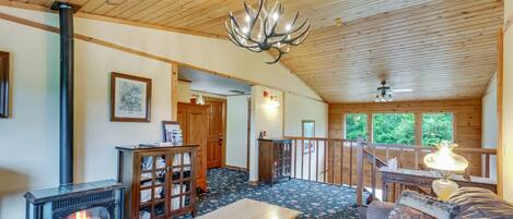 A cozy cabin interior with a wood-burning stove, a cushioned sofa, a wooden table, and a chandelier.