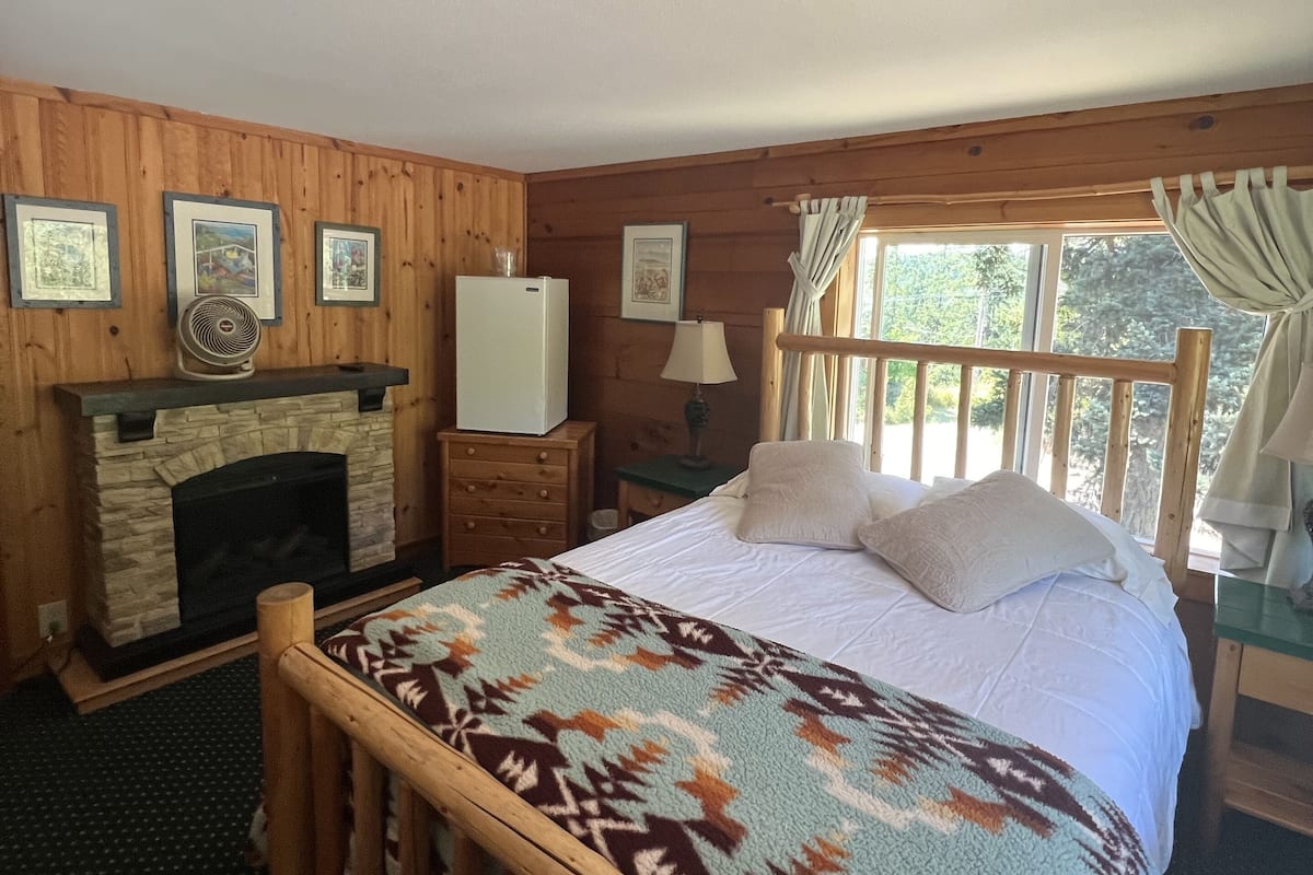 A wooden cabin bedroom with a stone fireplace, a bed with a patterned blanket, and framed pictures on the wall.