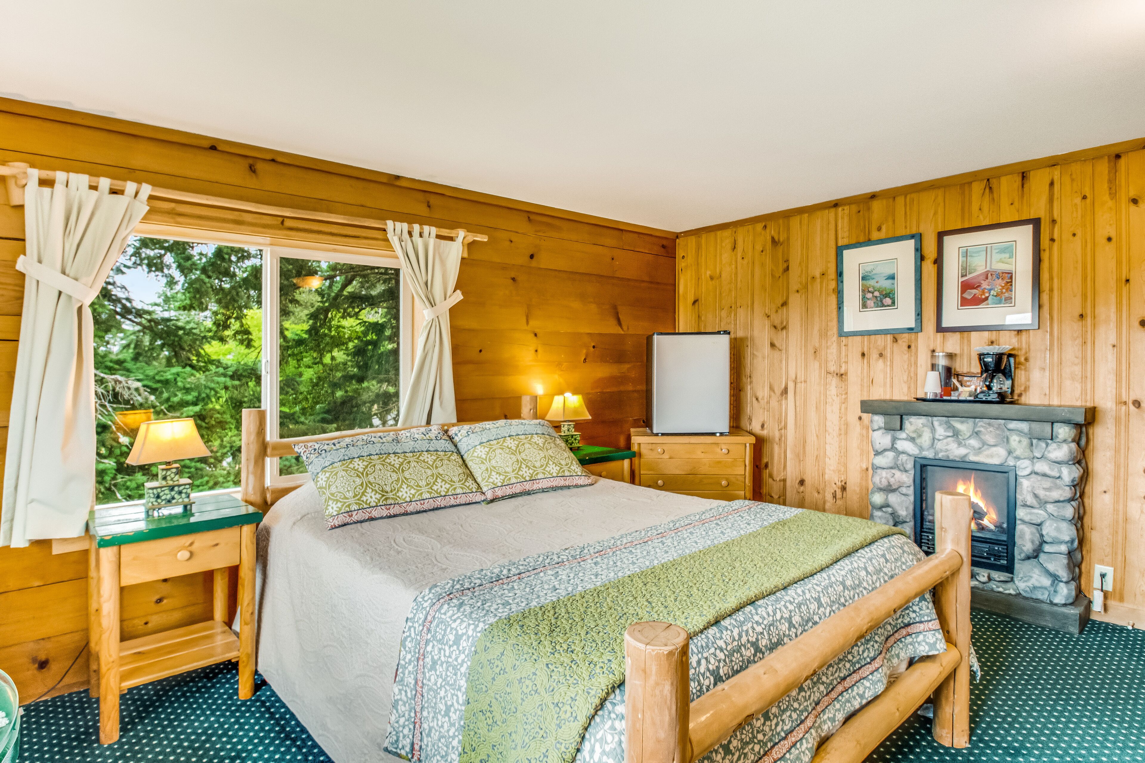 A cozy wooden cabin bedroom with a bed, nightstands, a fireplace, and a window with curtains.