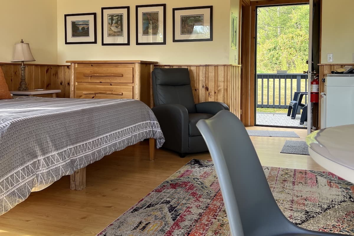 A cozy bedroom with a bed, a chair, and a small table. There are framed pictures on the wall and a window with a view of the outdoors.