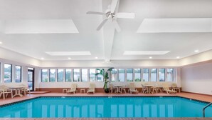 An indoor swimming pool with clear blue water, surrounded by lounge chairs and tables, and large windows providing views of the outside. - Wingate by Wyndham West Monroe (West Monroe)