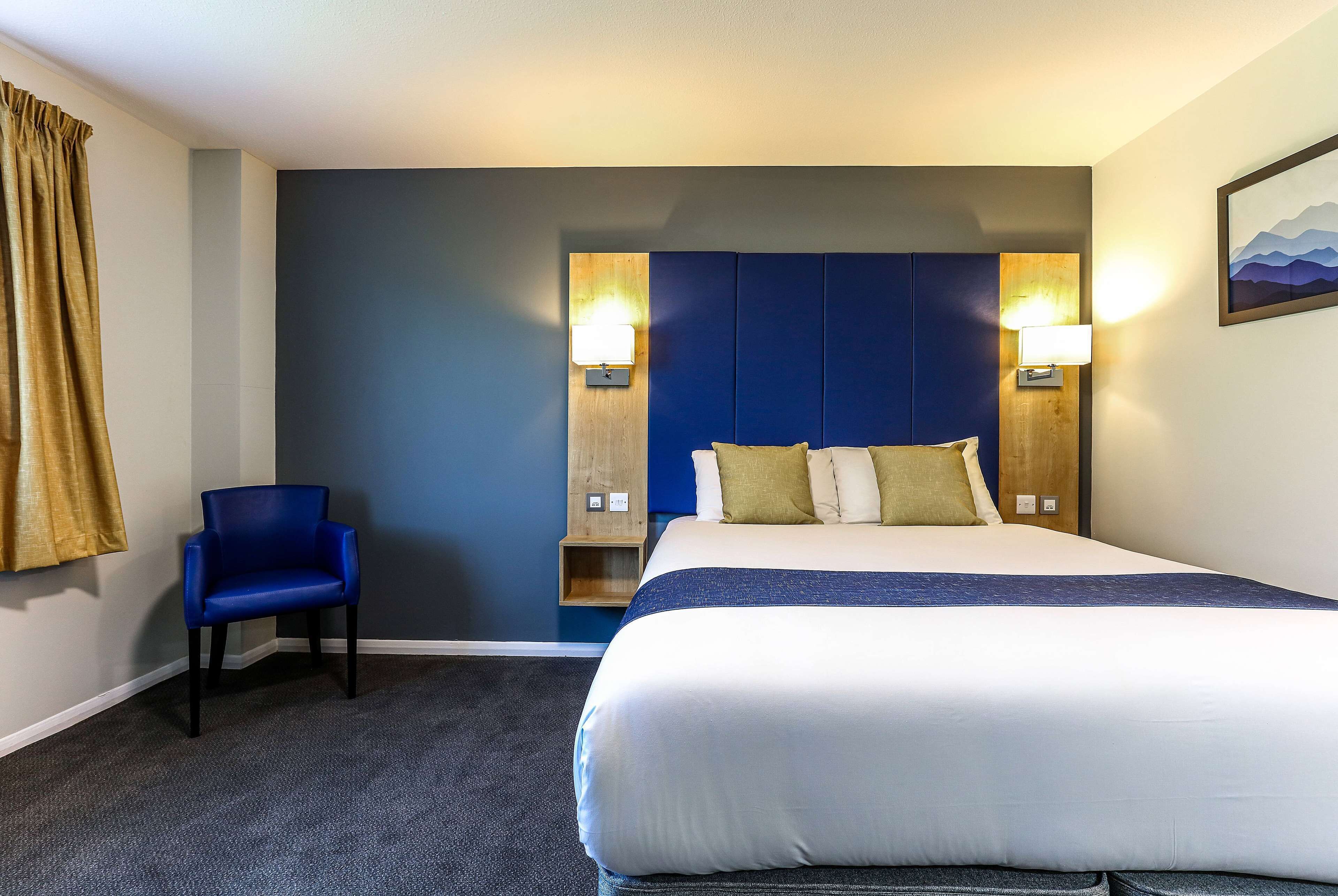 A hotel room with a large bed, a blue chair, and two wall-mounted lamps.