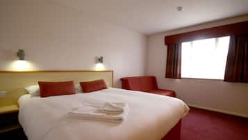 Habitación de hotel con una cama grande, dos almohadas, una silla, una ventana con cortinas y una lámpara de pared.