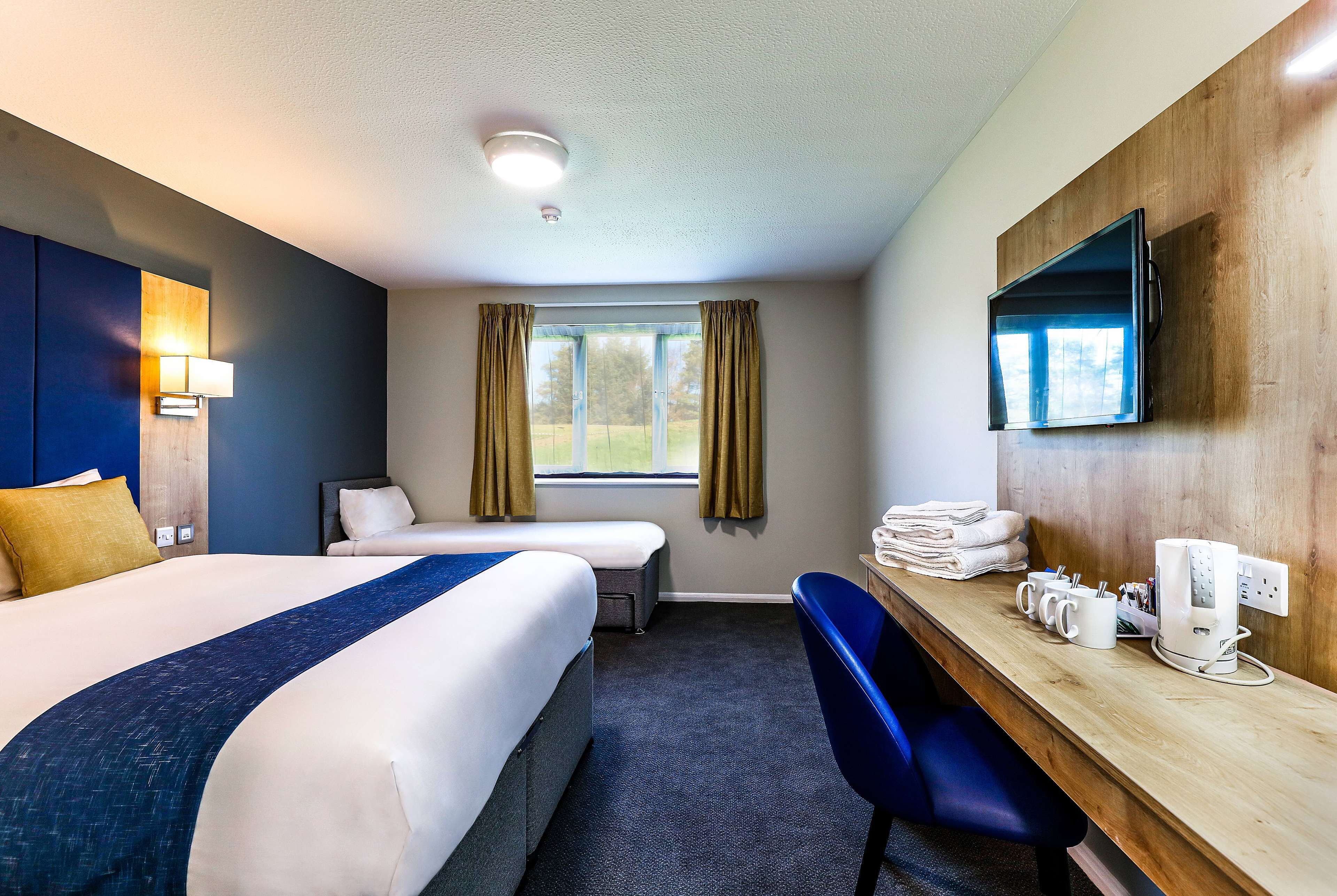 A hotel room with two beds, a desk with a chair, a TV, and a window with curtains.