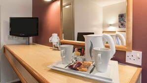 A hotel room with a wooden counter, a television, a mirror, and a tray with a mug, spoon, and a packet of snacks.