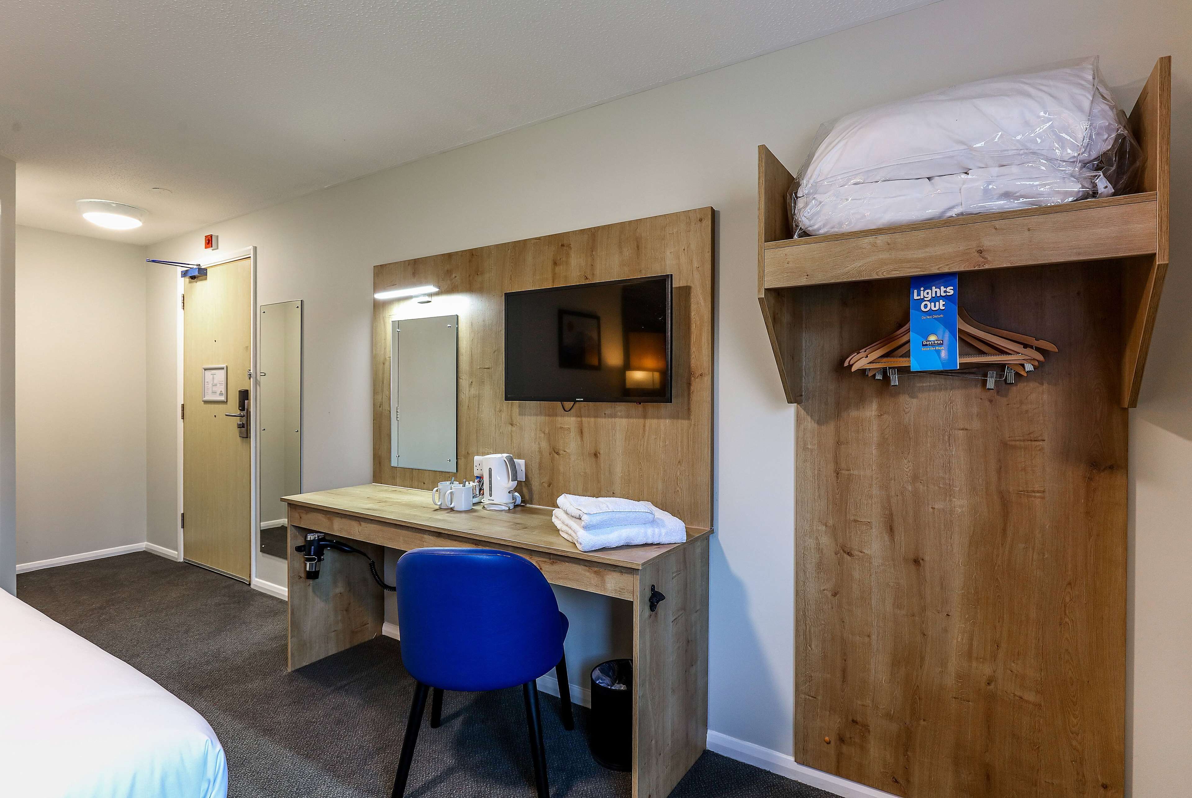 A hotel room with a wooden desk, a blue chair, a bed, a mirror, a television, and a shelf with a 'Lights Out' sign.