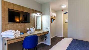 Habitación de hotel con un escritorio de madera, una silla azul, una cama con colchón azul, un televisor montado en la pared y un pequeño estante con toallas.