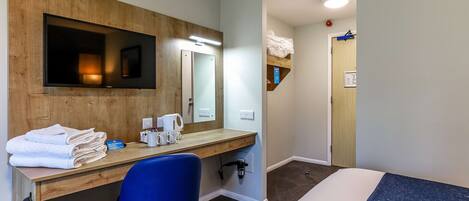 A hotel room with a wooden desk, a blue chair, a bed with a blue mattress, a TV mounted on the wall, and a small shelf with towels.