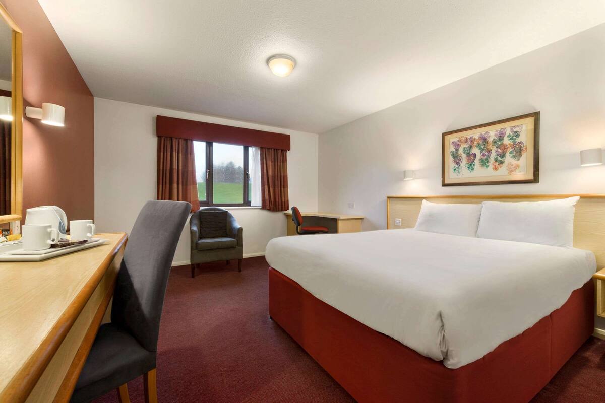 A hotel room with a large bed, a desk with a chair, a small table with cups, a window with curtains, and a framed picture on the wall.