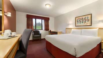 A hotel room with a large bed, a desk with a chair, a small table with cups, a window with curtains, and a framed picture on the wall.
