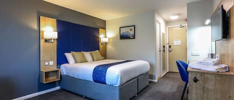 A hotel room with a bed, a desk, a chair, a wall-mounted TV, and a door leading to another room.