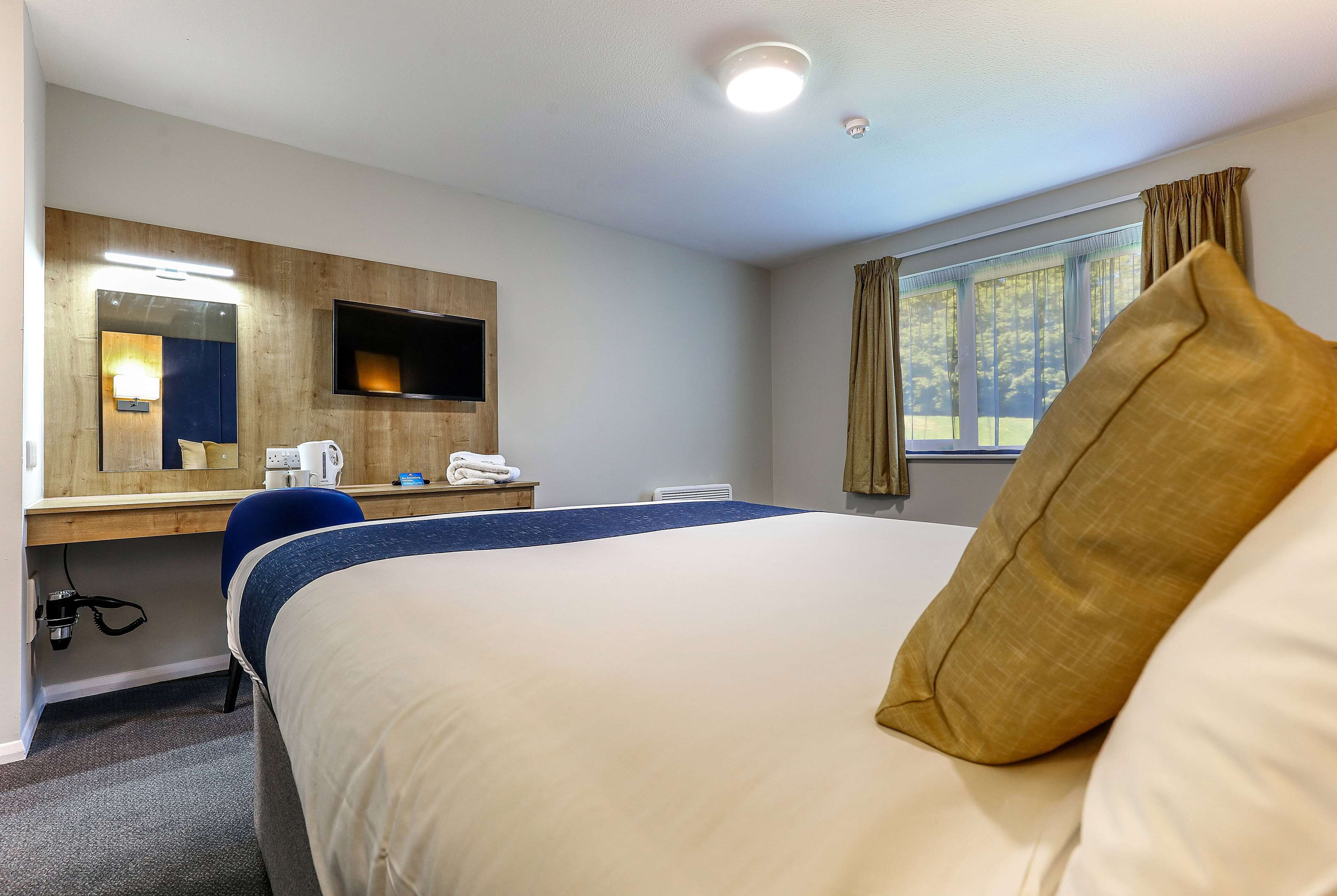 A hotel room with a bed, a desk with a computer, a mirror, and a window with curtains.