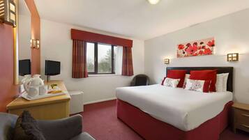 A hotel room with a large bed, a desk with a computer, a chair, and a window with curtains.