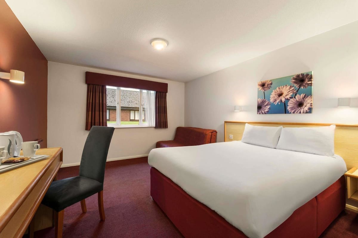A hotel room with a bed, a desk with a chair, a window with curtains, a painting on the wall, and a lamp.