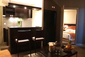 Quarto de hotel pequeno com cozinha compacta, área de refeições com duas cadeiras, uma mesinha com cesta de frutas e champanhe, e uma cama visível através de uma porta aberta.