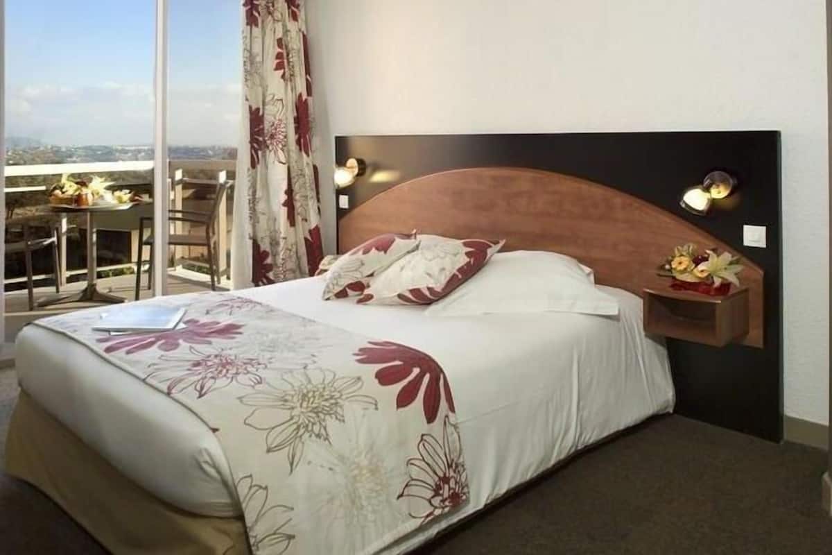 A hotel room with a bed, a nightstand, a window with curtains, and a view of the city.