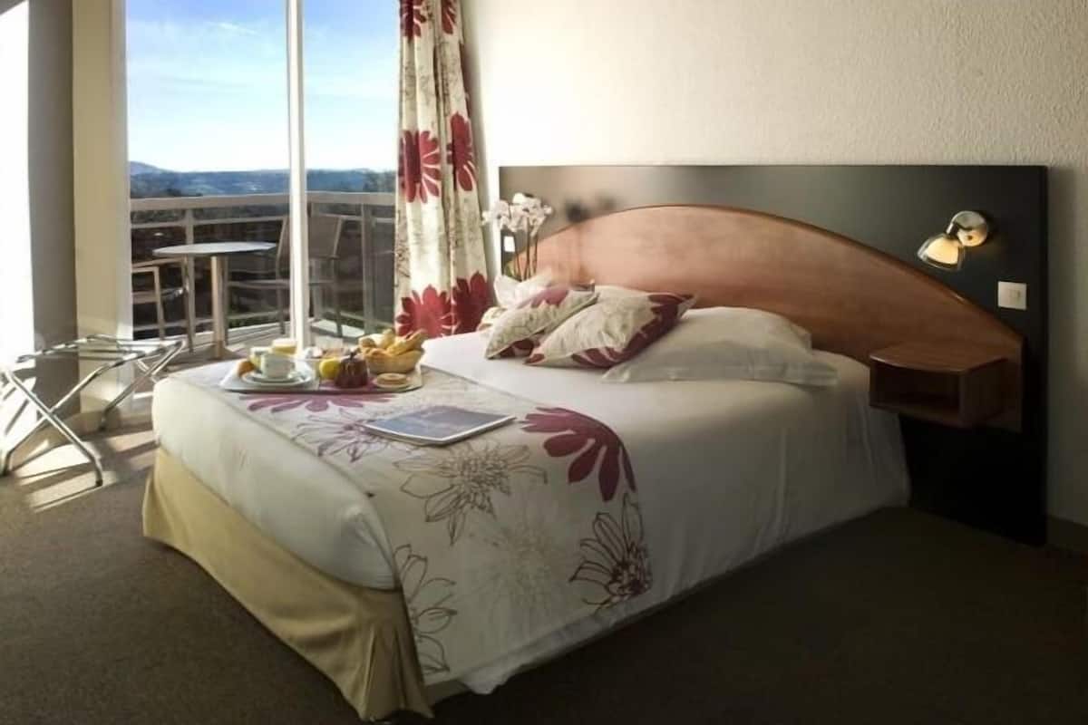 A hotel room with a large bed, a small table with a chair, a window with a view, and a nightstand with a lamp.