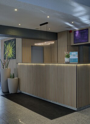 A reception desk with vertical wood paneling, a mounted TV, and a potted plant. - Waldhotel Wandlitz (Wandlitz)