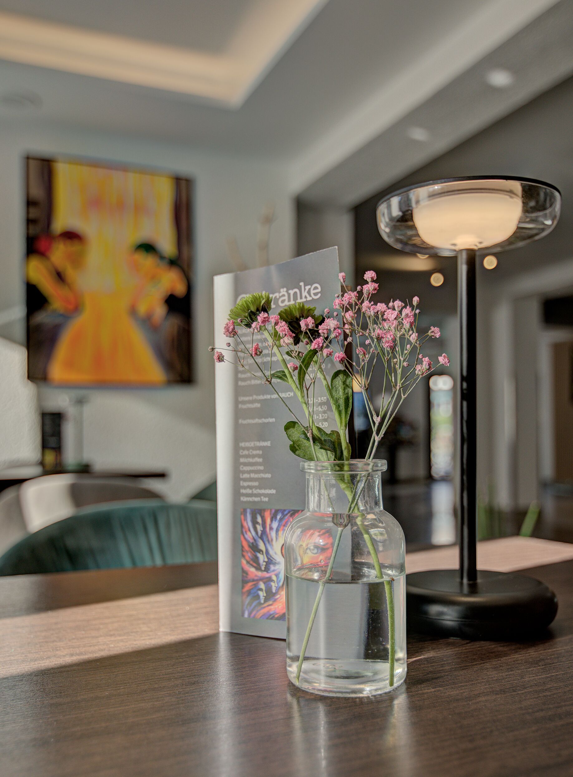 A clear glass vase with pink flowers on a wooden table, a menu, and a modern lamp.