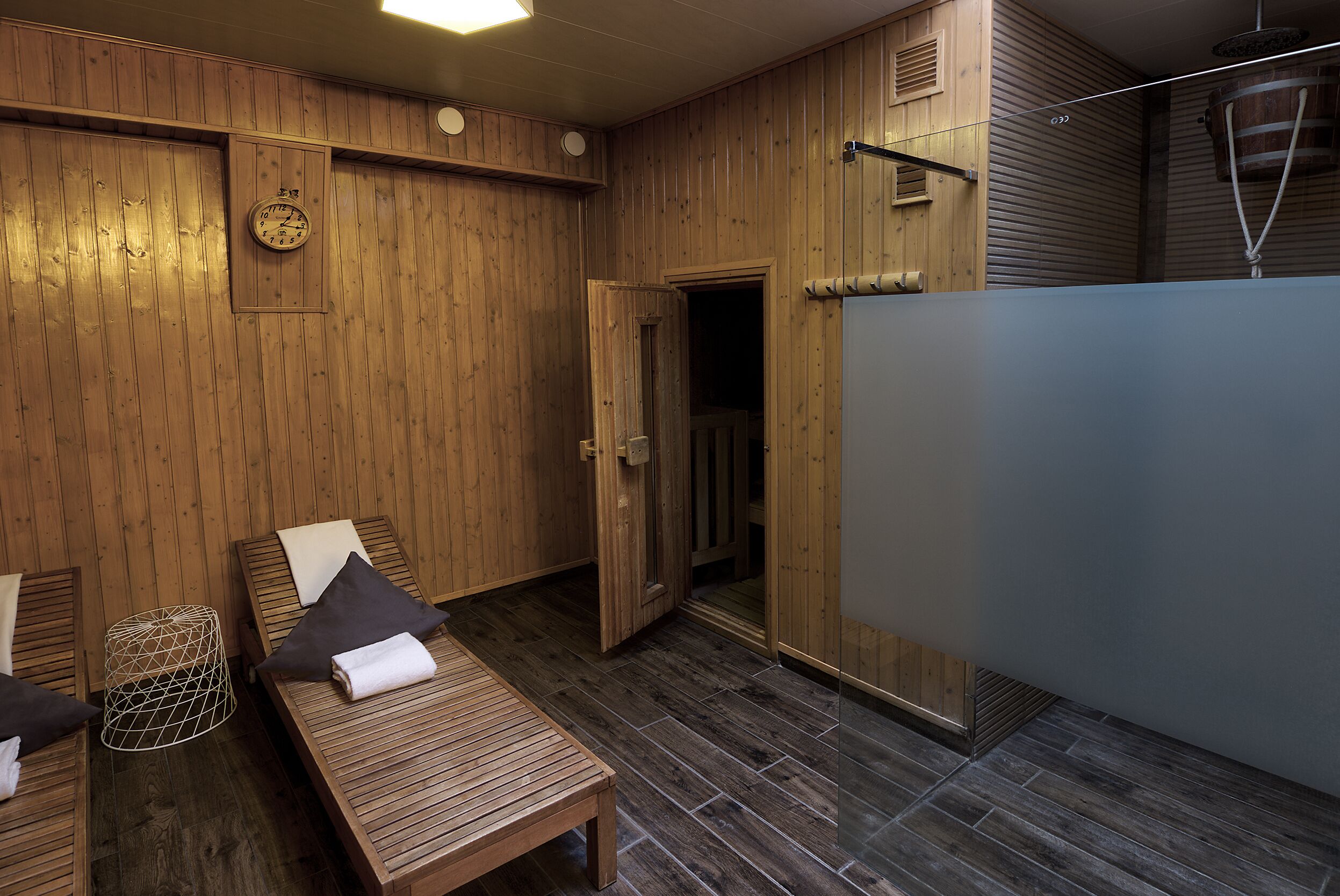 Una habitación de madera con una cama, un banco, un taburete pequeño y una puerta que da a otra estancia.