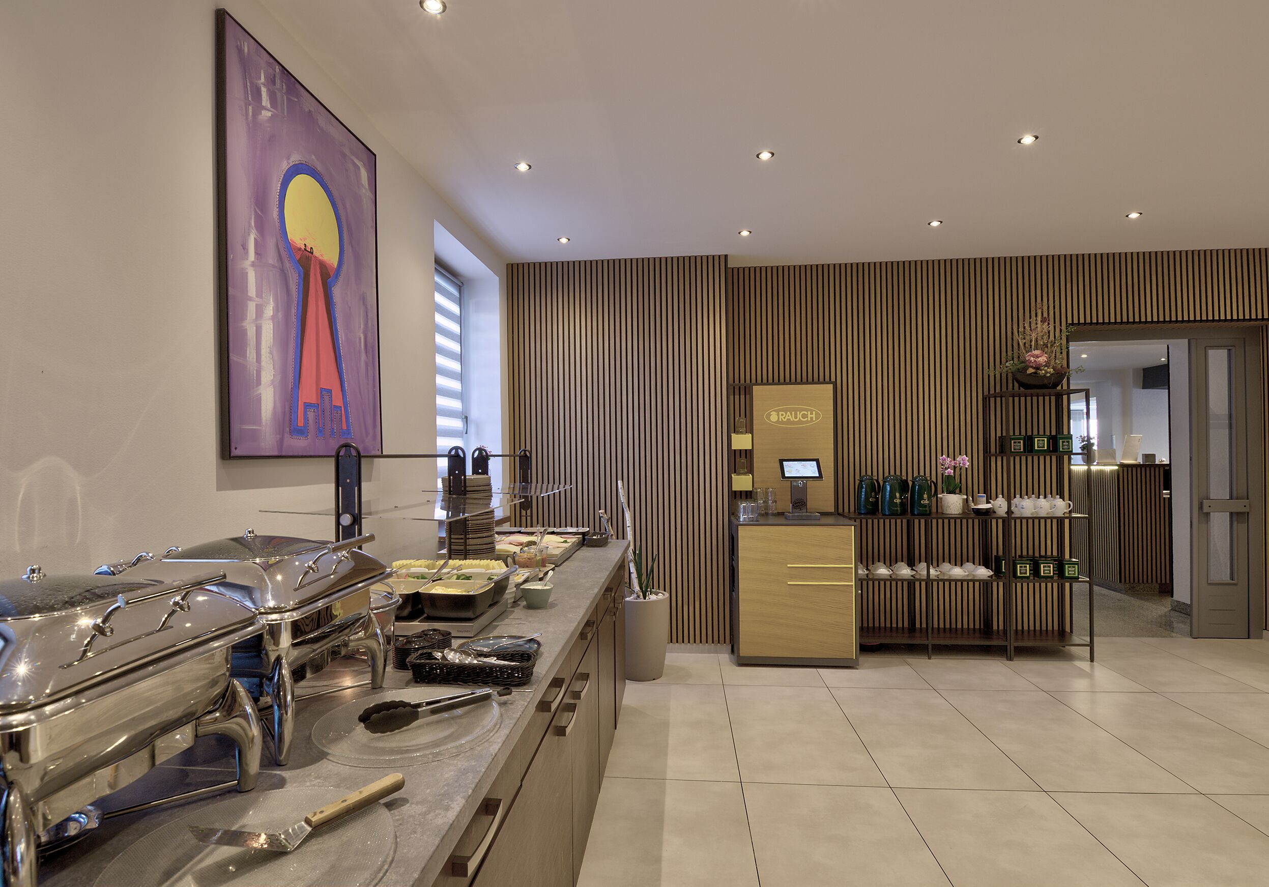 Zona de bufé con diversas opciones de comida, una estación de café, una estación de té y un mostrador de recepción con un monitor.