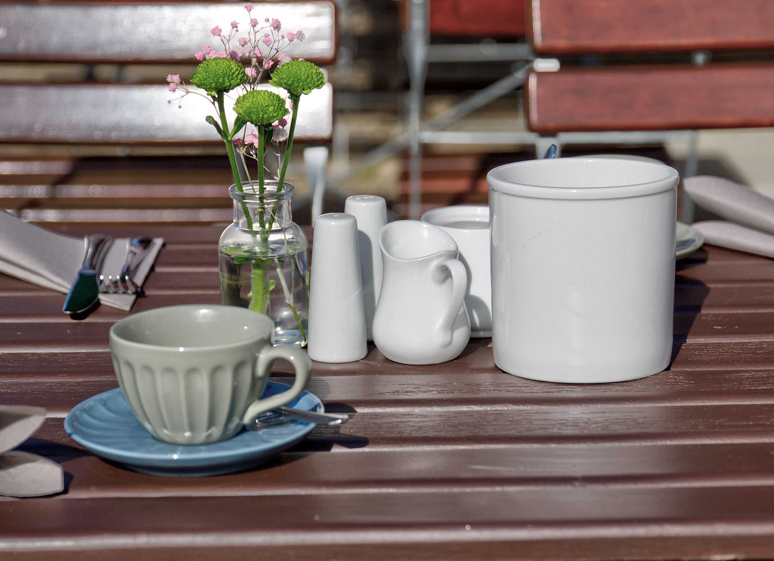 Una mesa con una taza blanca, una jarra blanca, un florero de vidrio transparente con flores verdes y un platillo azul.