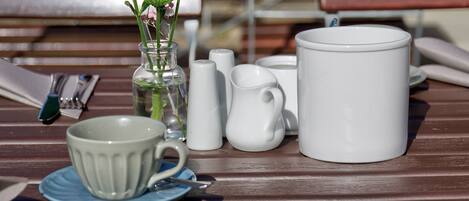 Una mesa con una taza blanca, una jarra blanca, un florero de vidrio transparente con flores verdes y un platillo azul.